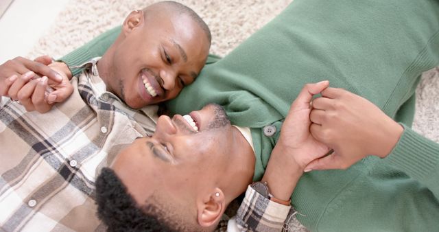 Happy diverse gay male couple lying on a carpet and embracing at home. Togetherness, relationship, love and domestic life, unaltered.