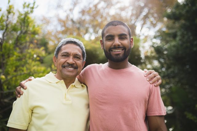 This image captures a joyful moment between a father and son standing together in a park. The father, wearing a yellow shirt, and the son, in a pink shirt, are smiling warmly at the camera. This photo can be used for family-oriented content, advertisements promoting family values, or articles about father-son relationships and bonding.