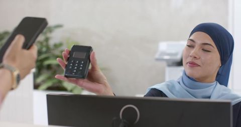 Receptionist wearing hijab holding contactless card reader while customer tapping smartphone