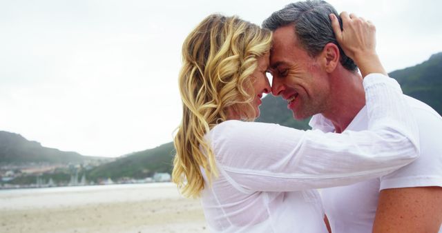 Couple embracing on beach, foreheads touching and smiling, setting a romantic and joyful mood. Perfect for use in advertisements, travel brochures, romance novels, and websites focusing on relationships and love. Includes an unidentifiable beautiful background, emphasizing a serene and affectionate moment.