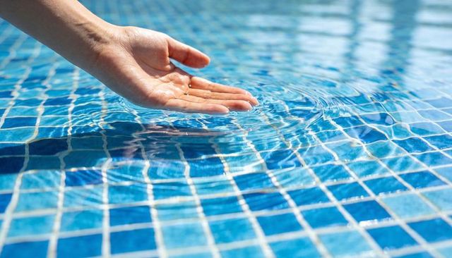 Hand touching pool water over blue mosaic tiles creating ripples and light reflections