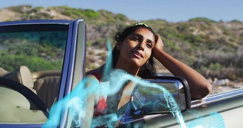 Leaning indian woman resting head in convertible by coastal dunes during sunlit roadtrip