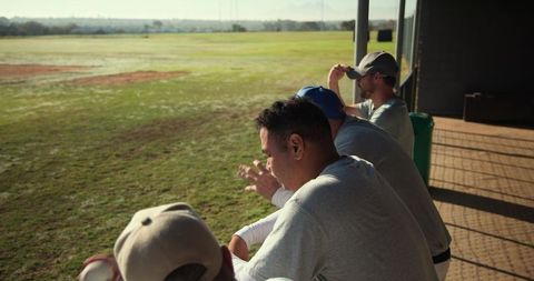 Baseball teammates watching outfield practice leaning on railing in sunlit training range