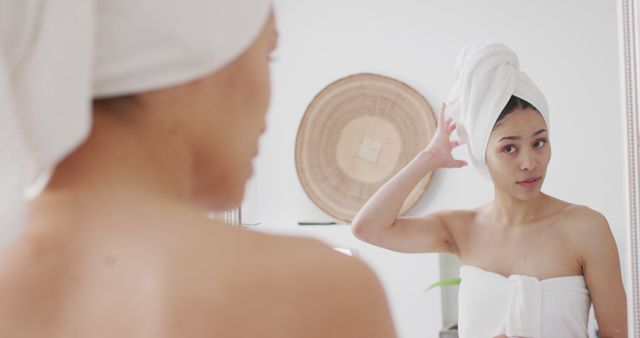 This photo depicts a woman in a bathroom adjusting a towel wrapped around her head in front of a mirror. Suitable for usage in wellness, self-care, hygiene, and lifestyle contexts. Ideal for articles, blogs, and advertisements focusing on self-care routines and home bathroom settings.