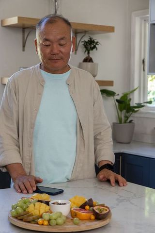 Senior asian man studying wooden fruit platter on marble kitchen counter with smartphone