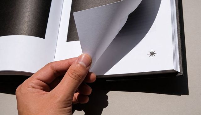 Hand turning page in hardcover book with star emblem, sunlit tabletop shadow, close-up