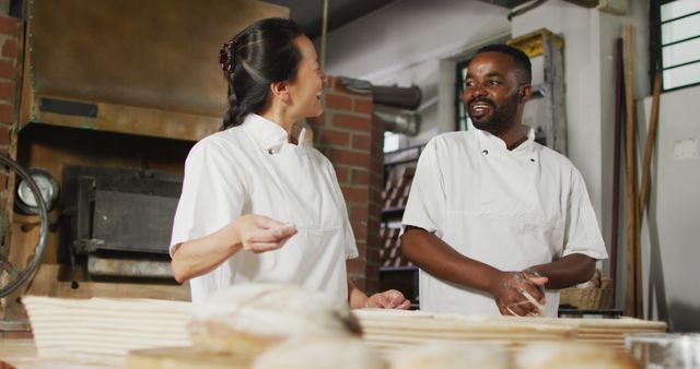 Two diverse bakery workers are communicating while preparing dough in a professional bakery. They are wearing white uniforms and appear to be enjoying their work. The background includes baking equipment and bread. This image is ideal for illustrating teamwork, culinary arts, and the bakery industry.