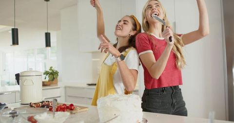 Joyful roommates singing with spatulas while frosting cake in bright modern kitchen
