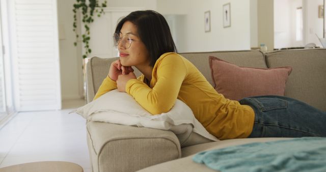 Woman is lounging on a comfortable sofa wearing a bright yellow sweater and stylish glasses, creating a cozy and happy atmosphere. Ideal for use in advertisements, lifestyle blogs, or social media posts about home comfort and interior design.