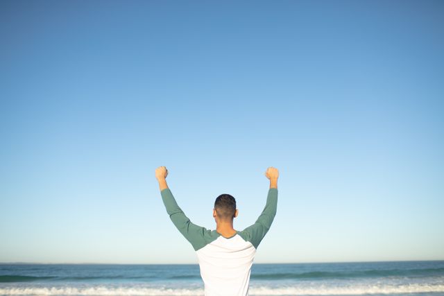 Man standing on beach with arms raised, celebrating success. Ideal for concepts of victory, freedom, and achievement. Perfect for motivational content, travel promotions, and lifestyle blogs.