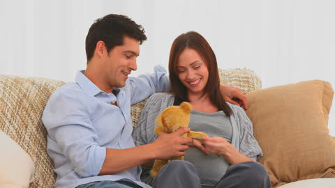Expectant Couple Playfully Engaging with Teddy Bear on Cozy Sofa