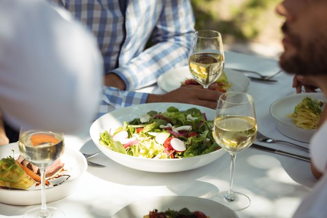 Close-up view of a dining table in a restaurant featuring a fresh salad, wine glasses filled with white wine, and various dishes. Ideal for use in content related to dining, gourmet cuisine, social gatherings, restaurant promotions, and lifestyle blogs.