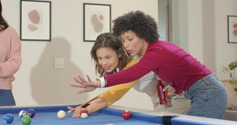 Diverse female friends leaning over blue-felt pool table, lining up shot while coaching