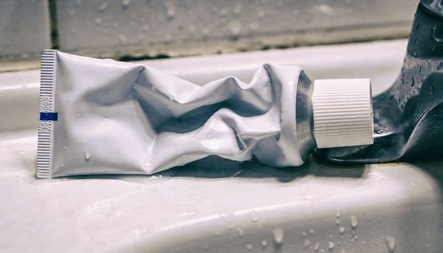Crushed white squeeze tube lying on wet ceramic sink ledge with cap and droplets