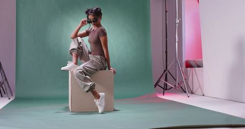 Edgy studio portrait of female model posing on cube, lifting shades, cargo pants