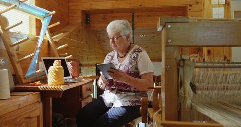 Senior woman weaving and using tablet in rustic workshop