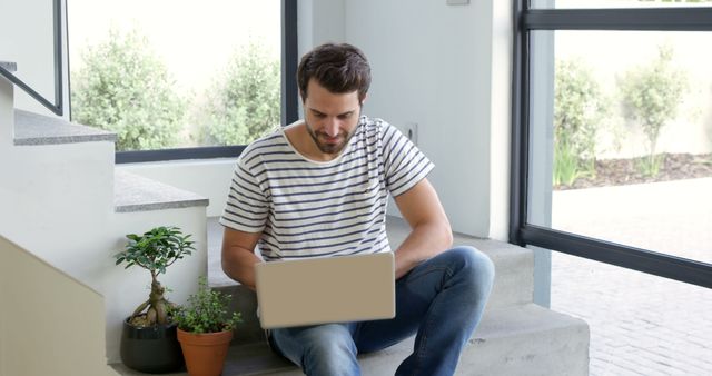 Young man seen in a relaxed home environment sitting on a staircase and using a laptop. The presence of indoor plants creates a pleasant and calm ambiance, making it perfect for themes of remote work, productivity, and home office setups. Ideal for articles on modern work-from-home trends, comfortable workspaces, and maintaining home-office balance.