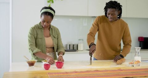 African american couple preparing pastry on kitchen island, filling ramekins, cutting dough