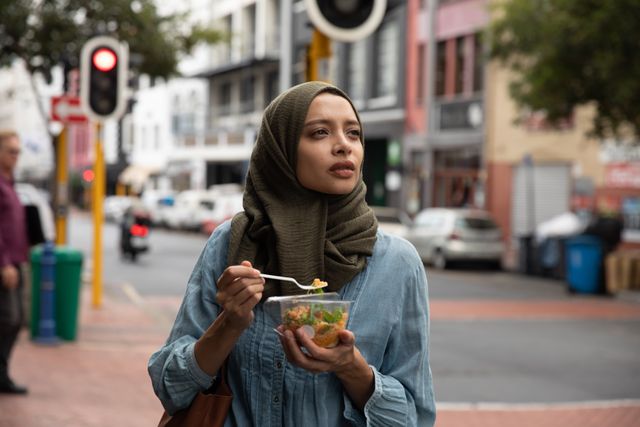 Biracial woman wearing hijab out and about on the go in the city, standing in street eating takeaway lunch holding bowl and fork. Commuter modern lifestyle.