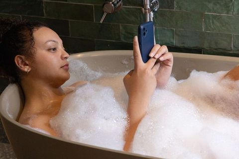 African american woman relaxing in bathtub scrolling smartphone amid bubbles and green tiles
