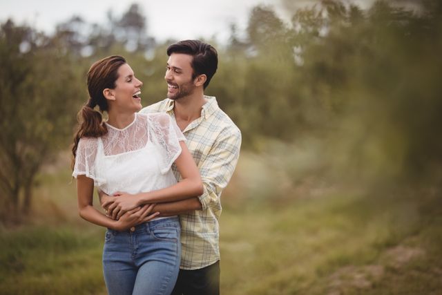 Young couple standing in an olive farm, embracing and smiling at each other. Ideal for use in advertisements, blogs, and social media posts about relationships, love, and outdoor activities. Perfect for promoting romantic getaways, countryside retreats, and lifestyle content.