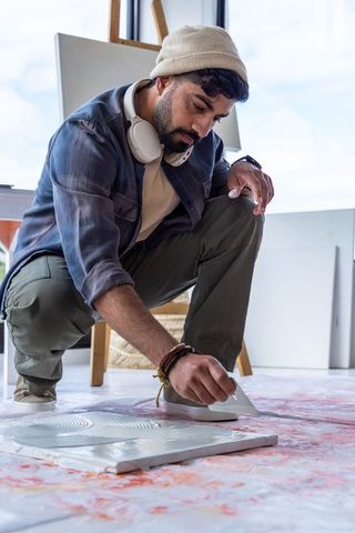 Indian artist crouching and scraping paint on floor canvas with palette knife