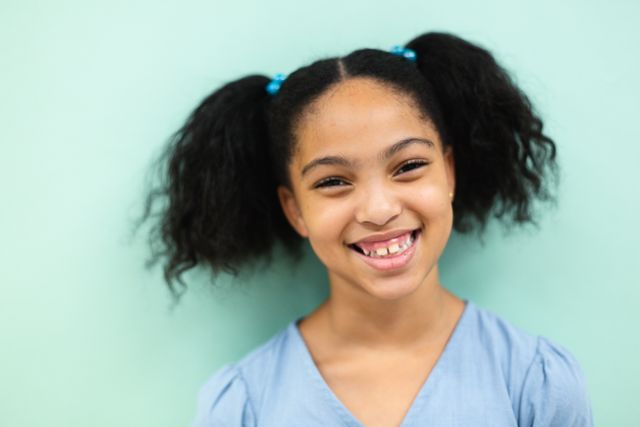 This image captures a joyful biracial elementary schoolgirl with pigtails smiling against a green background. Ideal for use in educational materials, school advertisements, childhood development articles, and promotional content for children's products or services.