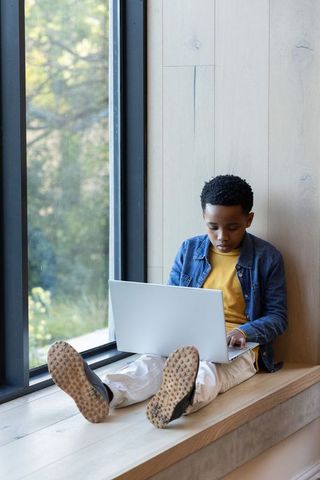 Curious african american boy studying on laptop by window bench in natural light
