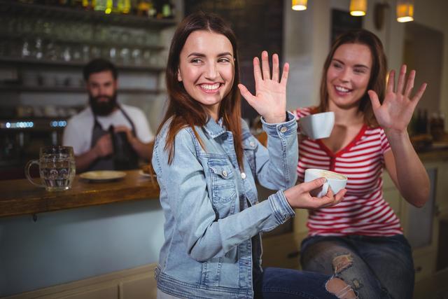 Two female friends are enjoying coffee in a cozy cafe, smiling and waving their hands. One is wearing a denim jacket, and the other is in a striped shirt. A barista is seen in the background. This image can be used for promoting social gatherings, coffee shops, friendship, and lifestyle blogs.