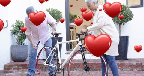 Mature couple adjusting white bicycle on porch with floating red hearts, romantic scene