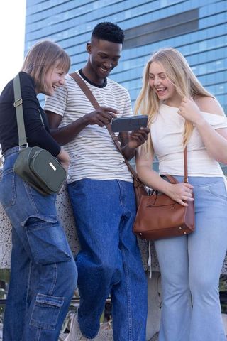 Friends laughing and sharing smartphone while leaning on concrete ledge in city
