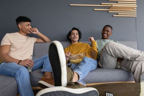Multiracial young men lounging on sofa sharing snacks and drinks during casual hangout