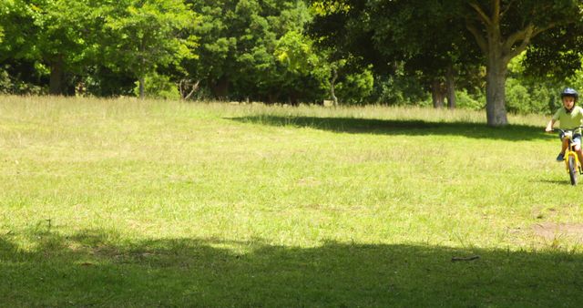 Child enjoying a sunny day while riding a bicycle in a shaded area of a park. Great for themes related to outdoor activities, childhood fun, healthy lifestyle, and nature. Useful for blogs, advertisements, and educational materials promoting physical activity and outdoor adventures for children.