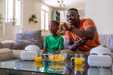 African American father guiding son playing video game on couch with VR headsets and snacks