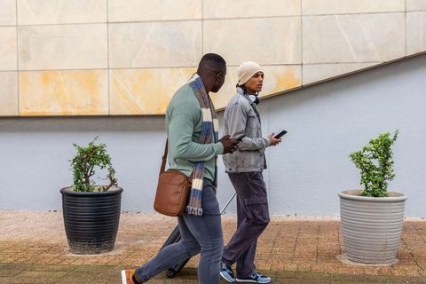 Two men walking with rolling suitcase and smartphones on city sidewalk by beige facade