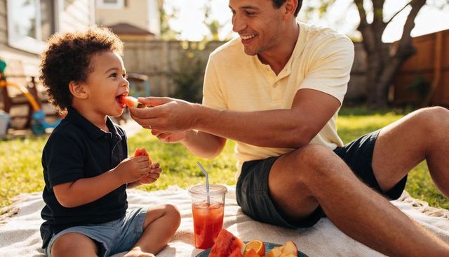 Dad feeding little boy watermelon on backyard picnic enjoying sunny family summer moment