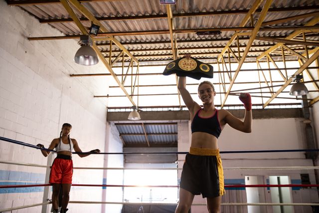 Biracial female boxers practicing in a boxing gym wearing sports clothes, one woman celebrating her victory holding belt up the other one standing in corner of ring. Strength sports achievement.
