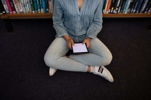 High angle view of a biracial female student wearing a jeans studying in a library, sitting on the floor, holding and using computer tablet.