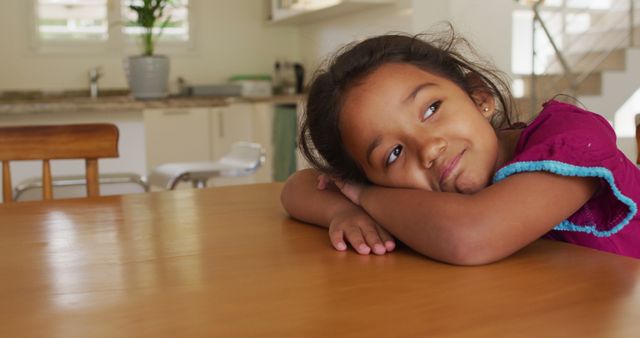 Young girl in a relaxed, happy moment, resting her arms on a wooden table. The bright, modern kitchen in the background suggests a warm, inviting home environment. Perfect for use in family lifestyle articles, advertisements for home-related products, or educational materials exploring child development and happiness.