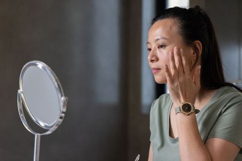 Mature woman applying skincare at vanity mirror, focusing on cheek and self-care routine