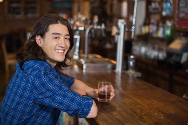 Young man with long hair smiling while holding a drink at a bar counter. Ideal for use in advertisements for bars, nightlife promotions, social media posts about leisure and relaxation, or articles about socializing and casual outings.
