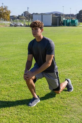Young man performing forward lunge on sunlit sports field for warmup and training