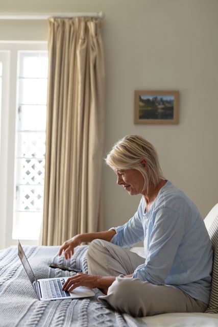 Mature woman sitting on bed using laptop, smiling and relaxed. Ideal for concepts related to senior lifestyle, technology use among older adults, home comfort, and independent living. Suitable for articles, advertisements, and blog posts about aging, remote communication, and domestic life.