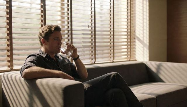 Man sipping water on modern sofa by sunlit slatted blinds, contemplative lounge moment