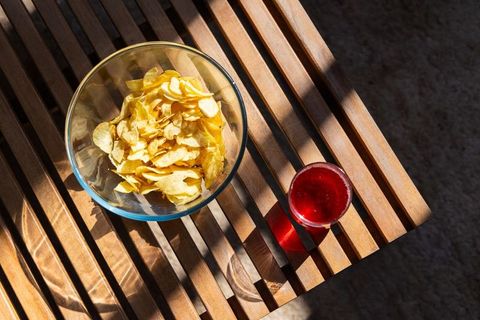 Sunlit potato chips sitting in glass bowl beside red drink on slatted wood table casting shadows