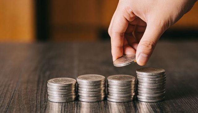 Hand placing coin on growing stacks of silver coins on dark wood table for savings