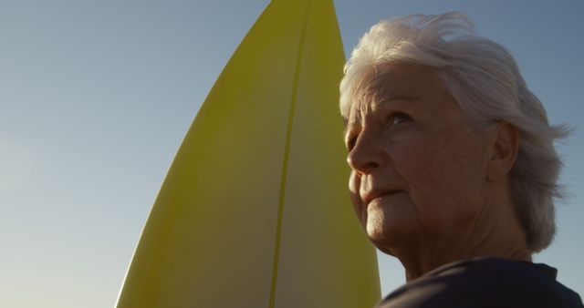 Senior woman standing with yellow surfboard near beach, looking contemplative at sunset. Perfect for themes related to active aging, fitness, outdoor activities, mental wellbeing, and inspiration. Can be used on websites, blogs, and advertisements promoting healthy lifestyles for seniors.