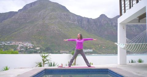 Woman practicing yoga warrior pose on poolside terrace with coastal mountain view