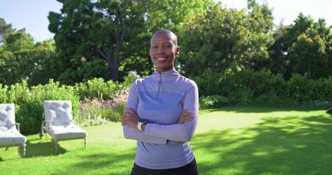Smiling woman standing with arms crossed in sunny backyard garden wearing lavender fitness top