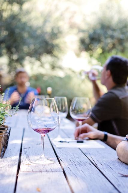 Friends enjoying wine tasting outdoors around rustic wooden table. Wine glasses and bottles are present, creating relaxed and enjoyable atmosphere. Perfect for advertisements related to wine, vineyards, leisure activities, or social gatherings. Suitable for illustrating outdoor dining, group activities, and casual social events.